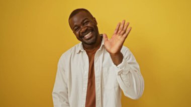 Genç adam, izole edilmiş sarı bir arka plana neşeyle el sallıyor. Çekici bir Afro-Amerikan erkeği gündelik bir ortamda pozitifliğini sergiliyor.