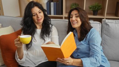 İki kadın keyifli bir anı paylaşıyor kitap okuyor ve sıcak bir oturma odasında kahvelerini yudumluyor. Ilık aydınlatma, aile sevgisini ve arkadaşlığını sergiliyor..