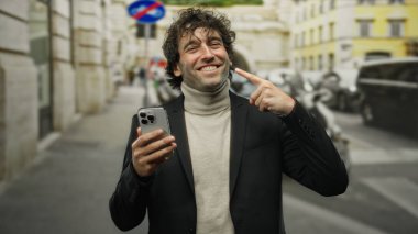 Balıkçı yaka ve spor ceketli mutlu bir adam şehrin dışında akıllı telefon kullanıyor, kendine güvenerek gülümsüyor ve neşeli bir ifadeyle kendini işaret ediyor..