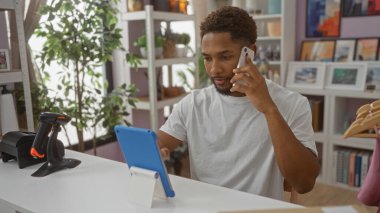 Genç adam, modern bir ev dekorasyonunda tablet kullanırken telefonla konuşuyor. Etrafı bitkilerle ve şık dekorasyonlarla çevrili.