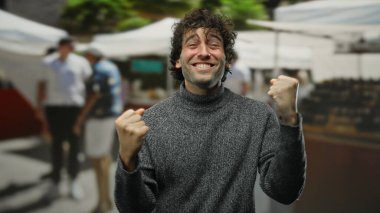 Genç adam hareketli bir şehir caddesinde özgüvenle gülümsüyor, yakışıklı ve çekici bir aura yayıyor açık hava piyasasının ortasında, neşeli ve pozitif titreşimleri somutlaştırıyor..