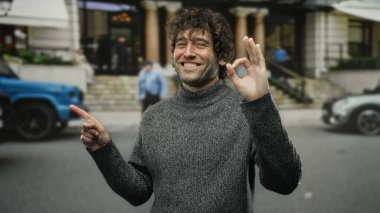 Genç İspanyol adam gülümsüyor ve el kol işareti yapıyor. Meşgul bir şehir caddesini işaret ederken açık bir günde şehir cazibesini rahat bir süveterle harmanlıyor..