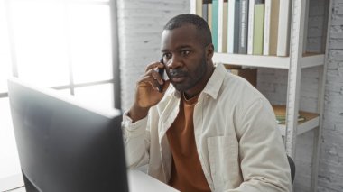 Ofisteki genç siyahi adam bilgisayar başında çalışırken telefonla konuşuyor, modern bir iş ortamı sergiliyor..