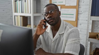 Modern ofis içinde cep telefonuyla konuşan genç adam, ciddi ifade ve düşünceli ruh halini yansıtıyor.