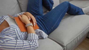 Evdeki bir kanepede rahat rahat uzanan genç bir kadın. Elleri oturma odasında görünür bir kitap tutuyor. Günlük giysiler giyiyor.