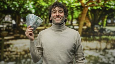 Parkta elinde bangladeş banknotlarıyla gülümseyen İspanyol adam, açık havada yeşil ağaçlarla güneşli bir günde mutluluğu ve finansal başarıyı gösteriyor..