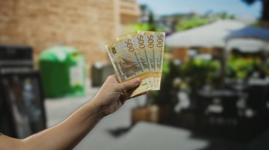 Elinde Norveç kronu banknotları tutan bir adam şehir caddesi terasında açık bir kafede masalarla çevrili ve insanlar güneşli gökyüzünün altında kahvenin keyfini çıkarıyorlar..