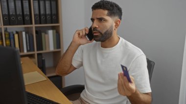 Ofis ortamında elinde pasaport ve kredi kartıyla konuşan genç adam endişeli görünüyor, arka planda kitaplık ve bilgisayarla içeride..