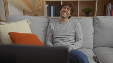 Genç adam rahat bir oturma odasında, modern dekorlu ve rahat koltuk minderleriyle televizyon izlerken rahatlıyor ve gülüyor..