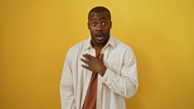 Genç, çekici, siyah, şaşırmış suratlı, günlük kıyafetler giyen, izole edilmiş sarı arka plan duvarına yaslanmış bir adam.