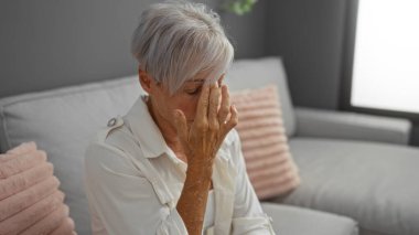 Gri mobilyalar ve pembe yastıklarla oturma odasında oturan kısa gri saçlı stresli orta yaşlı kadın.