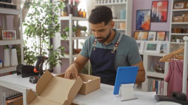 Genç adam bir ev dekorasyon mağazasında çalışıyor, eşyaları bir kutuda paketliyor, iç dekorasyon parçalarıyla çevrili ve görevleri yönetmek için bir tablet gibi modern teknoloji kullanıyor.