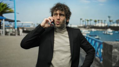 Genç İspanyol bir adam açık deniz manzaralı bir limanda teknelerle telefonda konuşuyor, rahat manzaraya güzelce karışıyor, yüzü odaklanmış ve özenli..