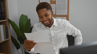 Yakışıklı, Afro-Amerikalı bir adam. Ofis ortamında evrak okurken gülümsüyor.