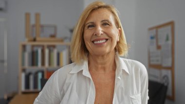 Ofis ortamında gülümseyen bir kadın, orta yaşlı, kısa sarı saçlı, arka planda beyaz gömlek, kitaplık ve pansiyon giyiyor.
