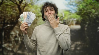 Parkta Güney Kore banknotlarını tutarken gülümseyen bir adam, açık havada güneşli bir günde yemyeşil ile çevrili finans sektöründe mutluluk ve başarı öneriyor..