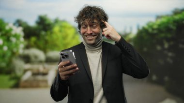 Genç adam elinde akıllı telefonuyla şakağını işaret ederken gülümsüyor. Arka planda bulanık siyah ceket giymiş yeşil bir parkta dikiliyor..