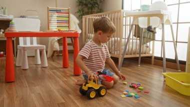 Kapalı bir anaokulu ortamında renkli oyuncaklarla oynayan sarışın çocuk, bir masa, sandalye, beşik ve ahşap bir zeminde sandalye var..