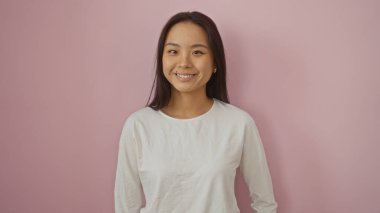 Güzel, genç Çinli bir kadın pembe arka planda poz veriyor, beyaz bir gömlekle çekici bir şekilde gülümsüyor..