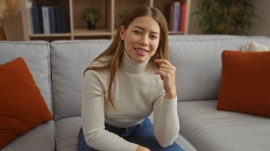 Modern dekorlu, rahat bir oturma odasında, kanepede oturan kadın beyaz bir kazak giyiyor ve sıcak bir şekilde gülümsüyor, rahatlatıcı ve davetkar bir atmosfer yaratıyor..