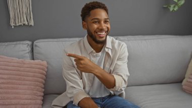 Yakışıklı genç adam gri dekorlu ve pembe yastıklı modern bir oturma odasında otururken gülümsüyor ve işaret ediyor.