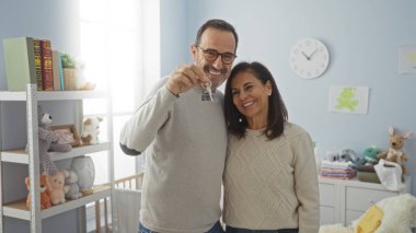 Orta yaşlı İspanyol bir çift oyuncak ve pastel dekorlarla dolu rahat bir bebek odasında anahtarlarını tutarken sevgi ve aileyi karşılama ortamında sembolize ediyorlar..