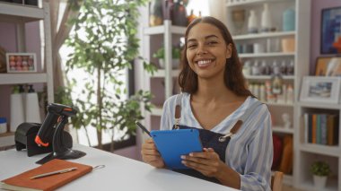 Bir ev dekoratörü veya dükkanda otururken elinde tabletle gülümseyen genç bir kadın, çeşitli dekorasyonlar ve ev bitkileriyle çevrili..