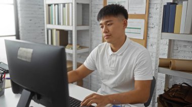 Evinde çalışan, bilgisayarlı bir masada oturan, modern, düzenli bir iç mekanda kitap raflarıyla çevrili genç bir adam.