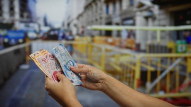 Kalabalık bir şehir caddesinde Meksika pezosu tutan adam, endüstriyel inşaat alanındaki finansal yatırımları ve kentsel iş atmosferini vurguluyor..