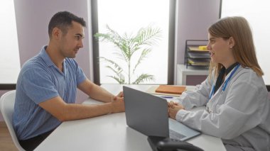 Klinikteki bir kadın ve erkek, masada steteskopla, profesyonel bir sağlık hizmetleri tartışmasına girişiyor, içeride doktor-hasta etkileşimini gösteriyor..