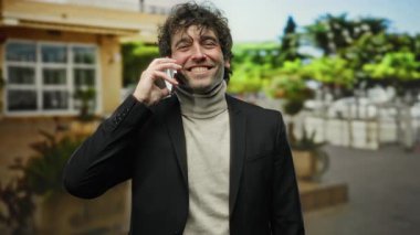 Ceketli genç adam arka planda canlı yeşillik olan bir şehir caddesinde cep telefonuyla konuşurken gülümsüyor.