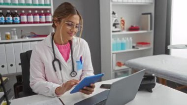 Klinikte çalışan bir kadın, düzenli bir tıp bürosundaki tablete bakar. Dosyaları, bilgisayarı, steteskopu ve raflarda şişeleri vardır. Profesyonel bir sağlık hizmeti ortamı sunar..