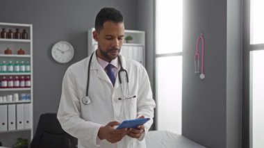 Steteskoplu İspanyol erkek doktor, modern hastane kliniğinde rafları ve tıbbi ekipmanları olan tableti inceliyor..