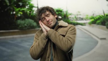 Genç İspanyol bir adam dışarıda, elleri yüzünün yanında, sıcak bir ceketle yeşil bir şehir arka planına karşı bir uyku jesti yapıyor..