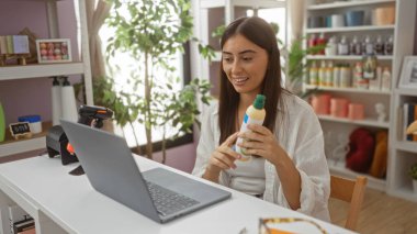 Kadın elinde bir ürün şişesi tutarken gülümsüyor ve arka planda raflar dolusu eşyayla bir dekor dükkanında dizüstü bilgisayar izliyor.