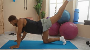 Spor salonundaki paspasın üstünde spor yapan genç bir adam. Arka planda spor topları ve ekipmanlar var.