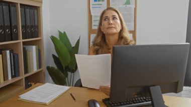 Ofiste çalışan kadın, arkaplanda dosyalar ve bitkilerle bilgisayar masasında belgeleri tutarken hayal kırıklığına uğramış gibi görünüyor.