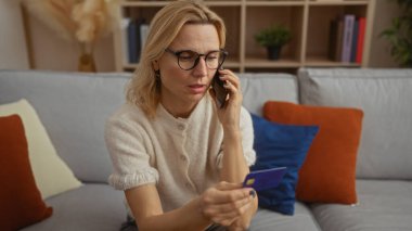 Evde elinde kredi kartıyla konuşan bir kadın, rahat bir oturma odasındaki şık bir koltuğa oturmuş, kapalı mekan iletişimi ve alışveriş anını yansıtıyor..