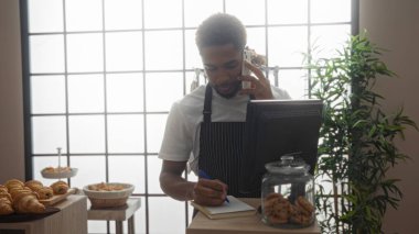 Fırın tezgahında çalışan genç adam, telefon siparişleri alıyor ve not defterine yazıyor, etrafında hamur işleri sergileniyor.