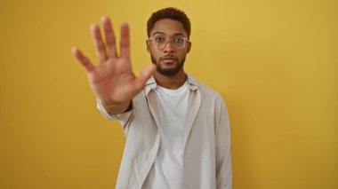 Genç adam elini sarı bir arka plana yaslayıp ciddi bir ifade sergilerken ve gündelik giysiler giyerken dur hareketi yapıyor..