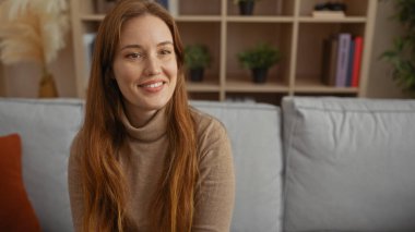 Kapalı bir oturma odasında modern dekorla gülümseyen kızıl saçlı kadın, rahatlama ve rahatlama için ideal sıcak ve davetkar bir atmosferi vurguluyor..