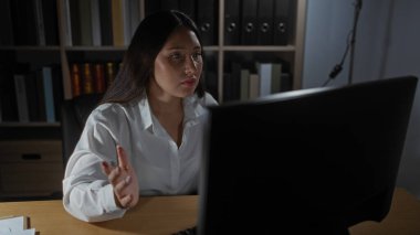 Ofiste çalışan genç, çekici, İspanyol bir kadın, arka planda raflar ve belgelerle dolu iyi düzenlenmiş bir iş yerindeki bilgisayar ekranına odaklanmış.