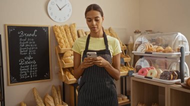 Fırında genç bir kadın akıllı telefonuyla etrafı ekmek ve pastalarla çevrili ve kapalı bir fırın tezgahında sergileniyor.