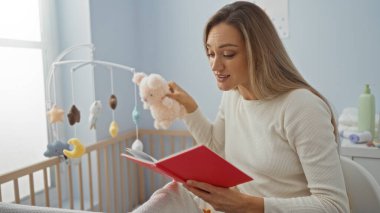 Bebek beşiğinin yanında oyuncak tutarken kitap okuyan bir kadın.