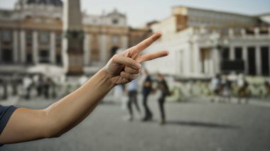 Arka planda bulanık turistler ve ikonik mimariyle St. Peter Meydanı, Vatikan 'da barış işareti yapan bir adamın eli..
