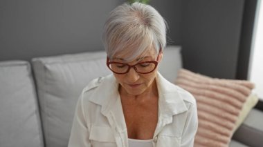 Gri saçlı ve gözlüklü olgun bir kadın modern bir oturma odasında rahat bir ifadeyle oturuyor.