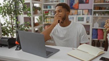 Delikanlı bir ev dekorasyonu dükkanında oturmuş arka planda yeşil ve dekoratif eşyalarla dolu bir masada otururken özenle dizüstü bilgisayara bakıyor.