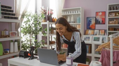 Güzel dekore edilmiş bir dükkanda çalışan, gülümseyen ve dizüstü bilgisayar kullanan genç bir kadın çeşitli ev dekorları ve yeşillikler arasında canlı ve canlı bir iç mekan ortamı sergiliyor.