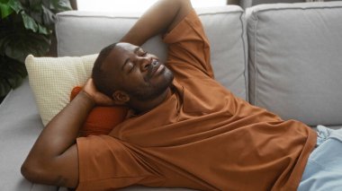 Modern bir oturma odasındaki rahat bir kanepede dinlenen genç adam, huzurlu ve memnun edici bir ifade sergiliyor.