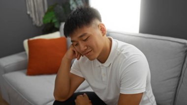 Modern bir oturma odasında gülümseyen yakışıklı genç Asyalı adam rahat bir iç mimarisi ve şık bir dekorasyonu var.